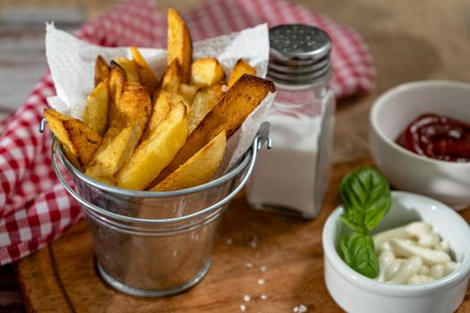French fries in a metal pot with aioli and ketchup on a wooden board.