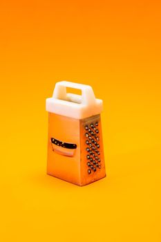 A small shiny metal grater isolated on orange background, close-up of cheese and food stainless grater.