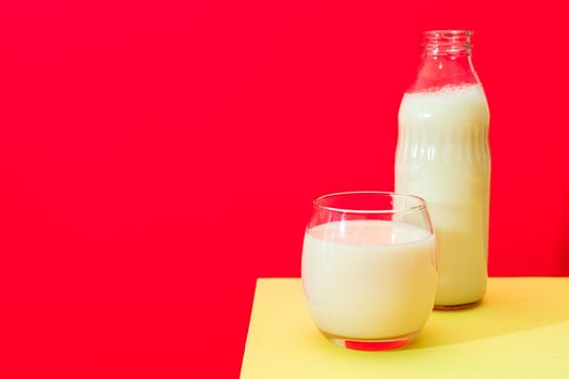 Glass bottle and big glass with milk on yellow table with red background. Copy space.