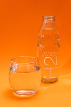 Bottle and glass tumbler with purified water on orange and light orange gradient background. Vertical Orientation