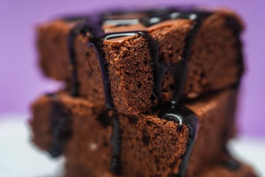 Very close shot of a Pile of Chocolate Brownies with chocolate strands falling on a white plate with purple background.