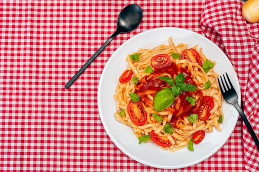 Top view of spaghetti pasta with a delicious homemade tomato sauce with homemade basil leaves served on a white plate on a red checkered tablecloth. Copy space. Natural and homemade food concept.