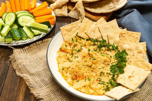 High view of a bowl with homemade Hummus with pita bread and crackers and in another bowl carrots and other vegetables. Concept of fresh, healthy and natural food, eaten at home.