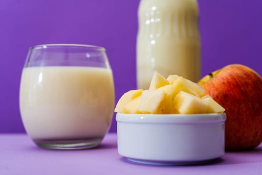 Glass bottle and large glass with milk and a whole red apple next to a bowl with apple cubes in a purple or violet environment. Copy space.