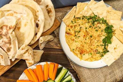 Aerial view of a bowl with homemade Hummus with pita bread and crackers and in another bowl carrots and other vegetables. Concept of fresh, healthy and natural food, eaten at home.