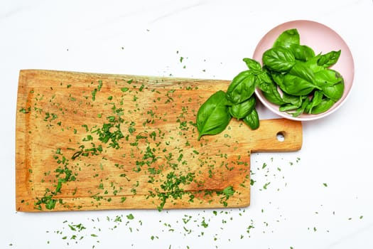 Fresh basil leaves in a small bowl next to a wooden cutting board on a white surface. Fresh, healthy and natural food concept. Copy space.