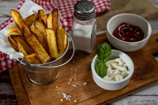 French fries in a metal pot with aioli and ketchup on a wooden board. High view.