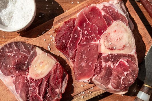 Two steaks of raw beef ossobuco on a wooden board with seasoning to flavour the pieces of meat. Animal protein concept, cheap cuts of beef. Aereal view.