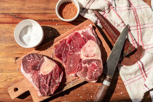 Two steaks of raw beef ossobuco on a wooden board with seasoning to flavour the pieces of meat. Animal protein concept, cheap cuts of beef. Aereal view.