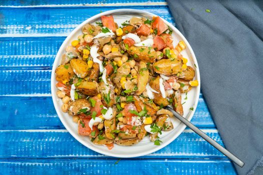 A delicious potato, chickpea, tomato and mushroom salad with parsley and aioli in a bowl on a blue rustic table. Healthy, homemade, vegan food. Top view.