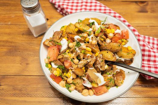 A delicious potato, chickpea, tomato and mushroom salad with parsley and aioli in a bowl on a wooden table. Healthy, homemade, vegan food. High view.