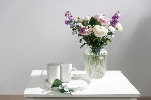 Bouquet of hackelia velutina, purple and white roses, small tea roses, matthiola incana and blue iris in glass vase is on the white coffee table with two tall cups for tea. Grey wall