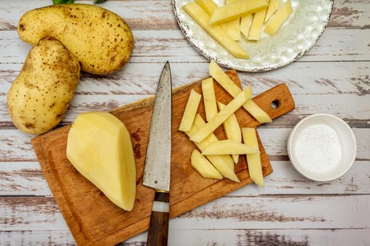 Wooden cutting board with a potato cut into a stick shape to make French fries. Half a peeled potato and a knife on a rustic table. Top view.