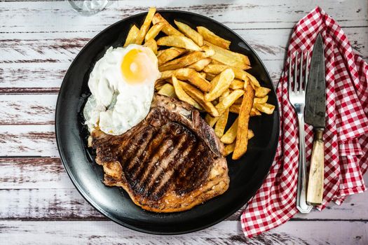 Plate with a juicy t-bone grilled or barbecued, accompanied by a portion of french fries and a fried egg. Top view. Traditional, homemade food concept.