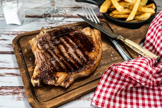 Incredible juicy t-bone cooked on the grill or barbecue on a wooden board accompanied by a french fries and with cutlery on the side. High view.