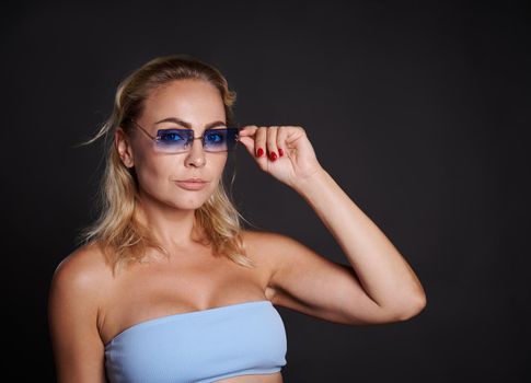 Confident portrait of a charming middle aged Caucasian woman with wavy blond hair wearing a blue top and blue sunglasses and looking at camera isolated over black background with copy ad space.