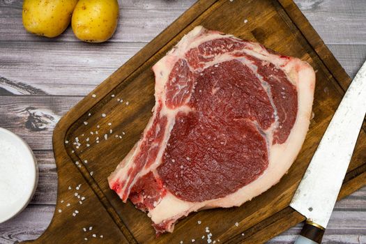 A bone-in steak or t-bone or porterhouse raw on a cutting board on a wooden table with one knfe and salt. Top view. High view. Close-up.