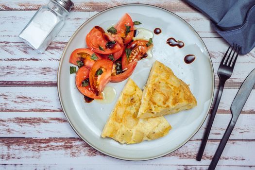 top view of a plate with two portions of Spanish potato omelette and sliced tomatoes with olive and sherry vinegar on a wooden rustic table.