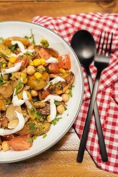 A delicious potato, chickpea, tomato and mushroom salad with parsley and aioli in a bowl on a wooden table. Healthy, homemade, vegan food. Partial view. vertical orientation.