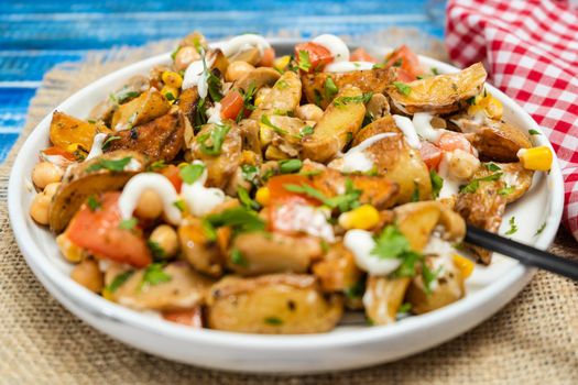 A delicious potato, chickpea, tomato and mushroom salad with parsley and aioli in a plate on a wooden table. Healthy, homemade, vegan food. Low view.