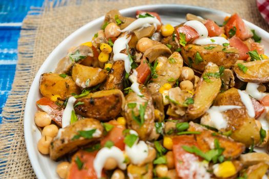 Close-up of A delicious potato, chickpea, tomato and mushroom salad with parsley and aioli in a bowl on a wooden table. Healthy, homemade, vegan food. Partial view.