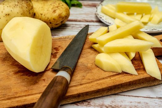 Wooden cutting board with a potato cut into a stick shape to make French fries. Half a peeled potato and a knife on a rustic table. High view.
