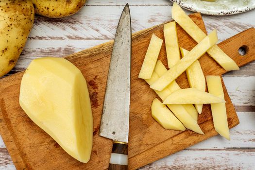 Wooden cutting board with a potato cut into a stick shape to make French fries. Half a peeled potato and a knife on a rustic table. Top view.