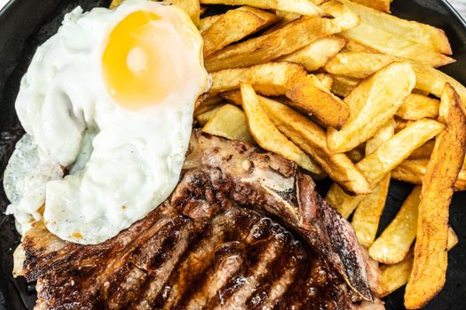 Plate with a t-bone grilled or barbecued, accompanied by a portion of french fries and a fried egg. Top view. Traditional, homemade food concept. Close up.