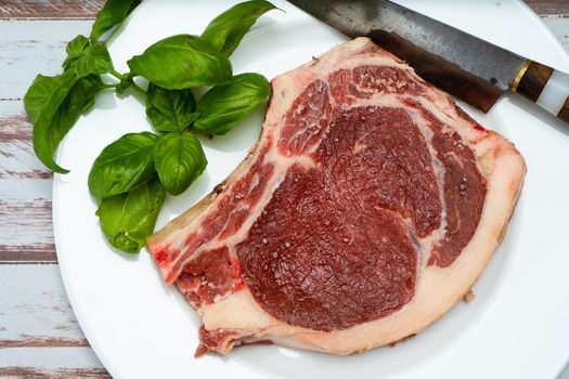 A bone-in steak or t-bone steak or porterhouse raw on a white plate on a wooden table with some green leaves. Top view. Horizontal orientation. Close-up.