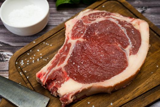 A bone-in steak or t-bone or porterhouse raw on a cutting board on a wooden table with one knfe and salt. Top view. High view. Close-up.