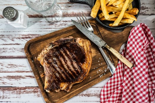 Incredible juicy t-bone cooked on the grill or barbecue on a wooden board accompanied by a french fries and with cutlery on the side. Top view.