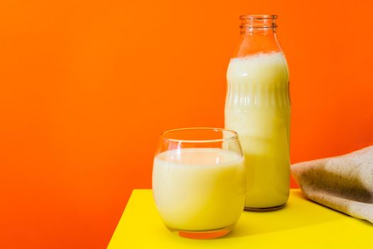 Glass bottle and large glass with milk on a yellow table with orange background. Copy space.