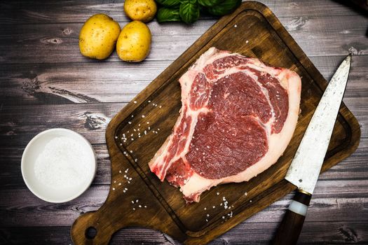 A bone-in steak or t-bone or porterhouse raw on a cutting board on a wooden table with one knfe and salt. Top view. High view. Close-up.