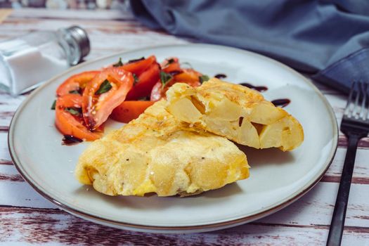 High view of a plate with two portions of Spanish potato omelette and sliced tomatoes with olive and sherry vinegar on a wooden rustic table.