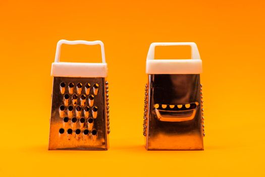 Two small shiny metal grater isolated on orange background, close-up of cheese and food stainless grater.