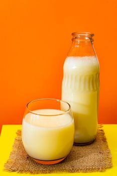 Glass bottle and big glass with milk on a square of rustic fabric on a yellow table with red background. Copy space. Vertical orientation