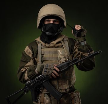 Russian soldier in military clothes with a Kalashnikov assault rifle on a dark background takes orders on a walkie-talkie