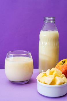 Glass bottle and large glass with milk and a whole red apple next to a bowl with apple cubes in a purple or violet environment. Copy space. Vertical orientation