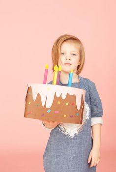 Portrait of an excited pretty little girl celebrating birthday and showing cake on over pink background.