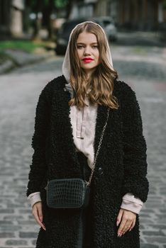 Fashion photo of young woman in black fur coat, jeanse and shoes at city street. beige jacket with a hood