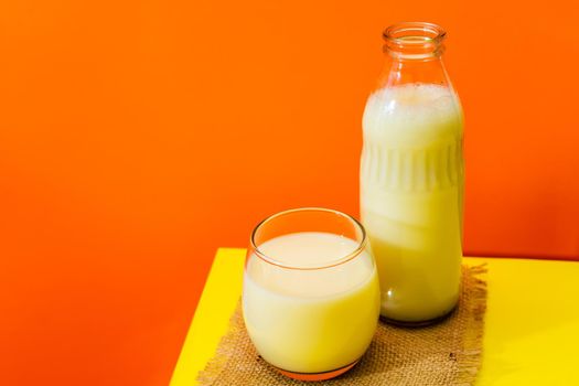 Glass bottle and big glass with milk on a square of rustic fabric on a yellow table with red background. Copy space. Horizontal orientation.