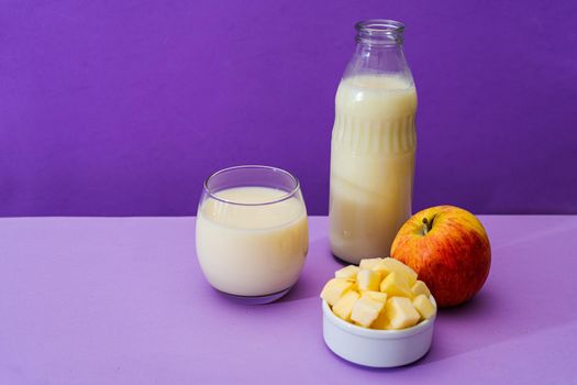 Glass bottle and large glass with milk and a whole red apple next to a bowl with apple cubes in a purple or violet environment. Copy space. Landscape orientation.