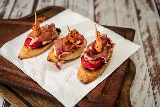 Bruschettas with serrano ham, cherry tomato and a grilled shrimp on a wooden board. Mediterranean food concept.