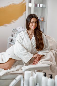 Woman in white bathrobe smiling at camera on daybed in spa center. Laser epilation concept