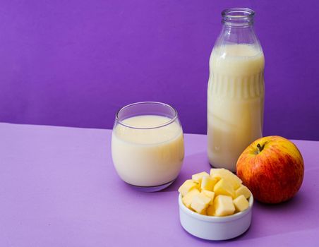 Glass bottle and large glass with milk and a whole red apple next to a bowl with apple cubes in a purple or violet environment. Copy space. Landscape orientation.