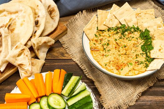 High view of a bowl with homemade Hummus with pita bread and crackers and in another bowl carrots and other vegetables. Fresh, healthy and natural food concept.