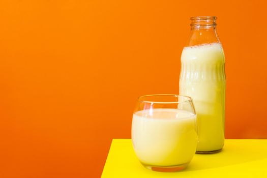 Glass bottle and large glass with milk on a yellow table with orange background. Copy space.