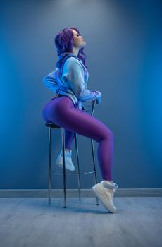 girl in stylish purple sportswear and with purple hair poses sexually on a bar stool