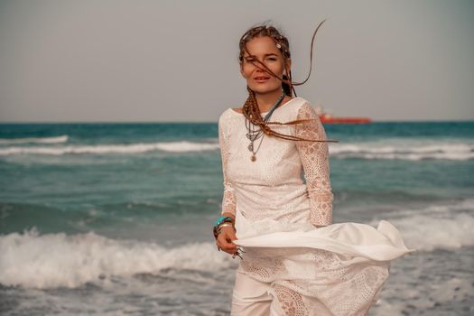 Model in boho style in a white long dress and silver jewelry on the beach. Her hair is braided, and there are many bracelets on her arms