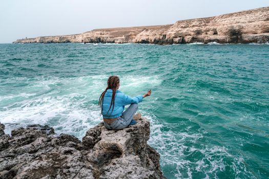 A woman in a blue jacket sits on a rock above a cliff above the sea, looking at the stormy ocean. Girl traveler rests, thinks, dreams, enjoys nature. Peace and calm landscape, windy weather